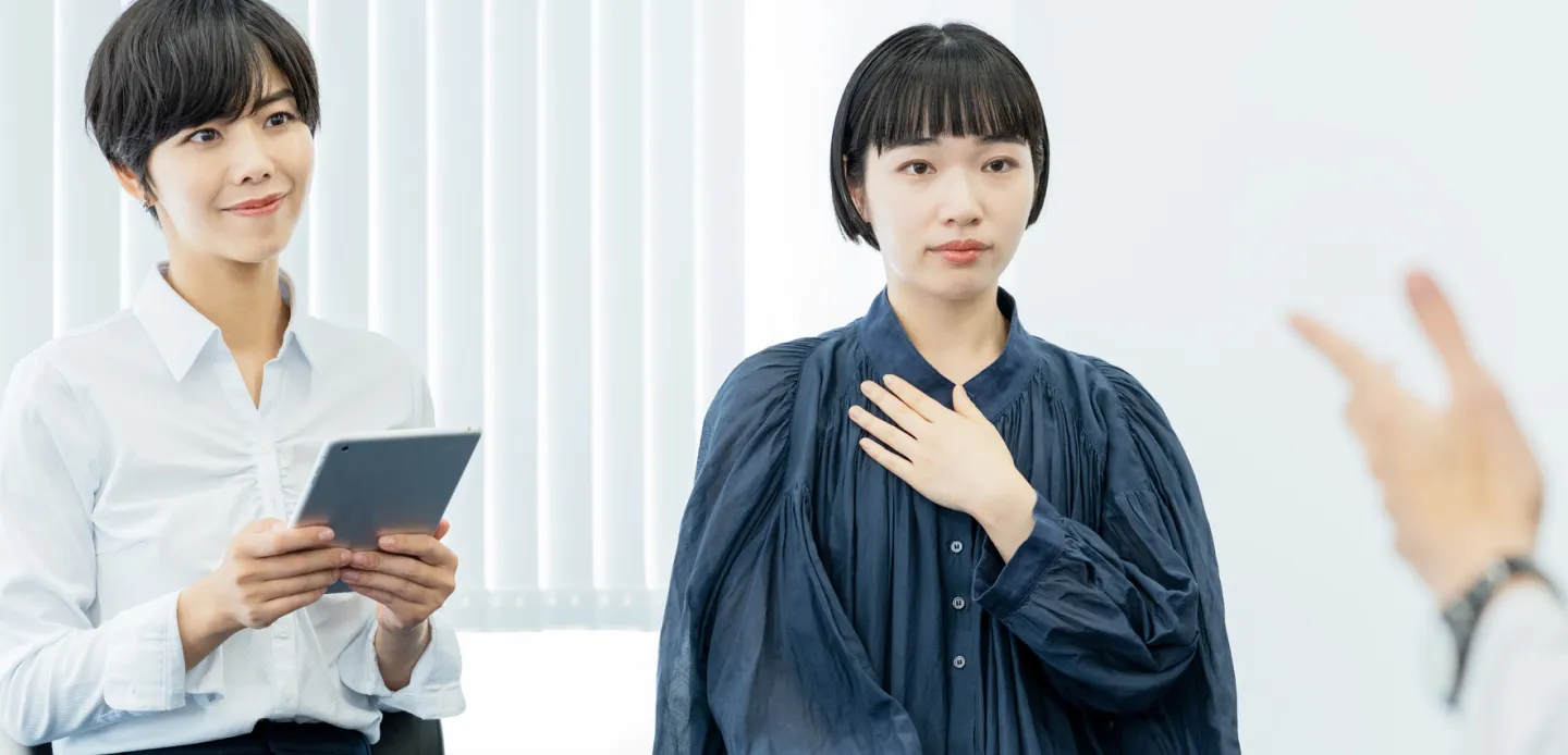 タブレットを持つ女性と胸に手を当てる女性が向かい合い、対話や面談をしている室内の様子