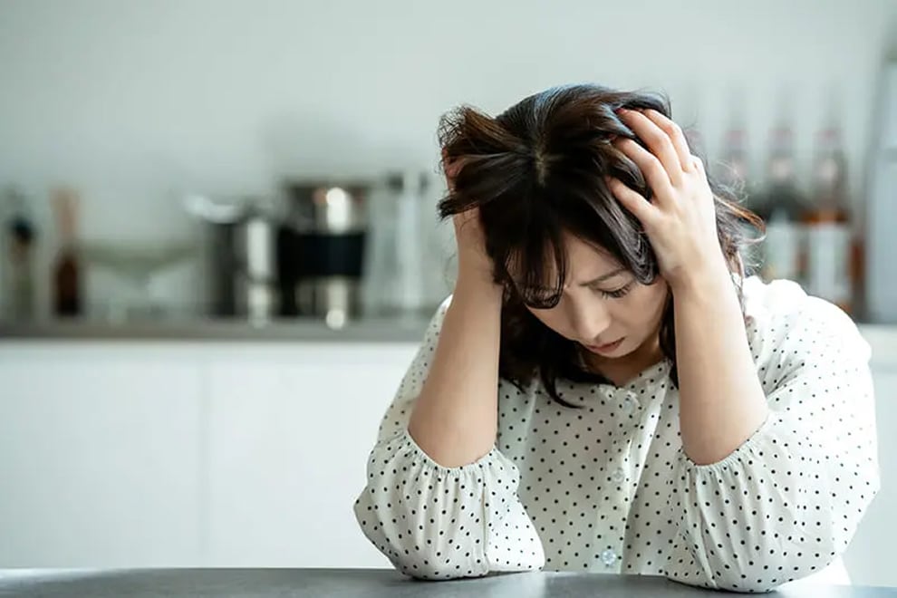 キッチンのテーブルに座り、両手で頭を抱え込みながら深刻な表情を浮かべる女性の様子