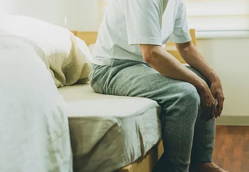 ベッドの端に腰掛け、前かがみの姿勢で両手を膝に置き静かに座る人物の様子